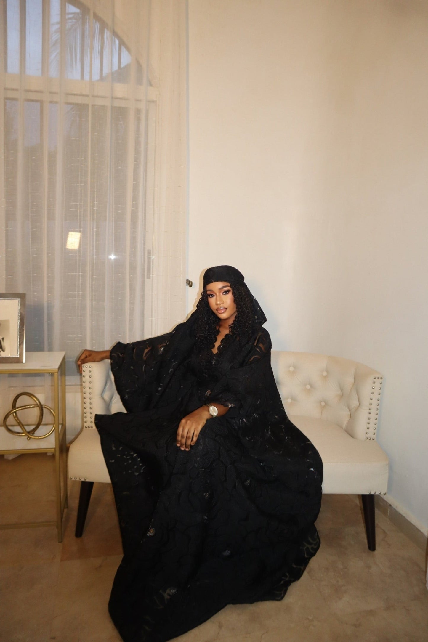 Woman in a black abaya sitting on a beige chair in a room with a window and light-colored walls.